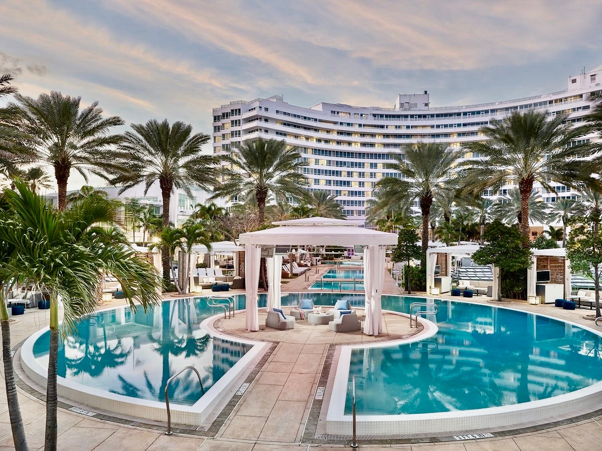The image captures multiple outdoor swimming pools surrounded by tall palm trees. Private cabanas are positioned along the poolside, creating serene lounging areas. The hotel structure looms in the background, featuring a modern design against a soft sky.