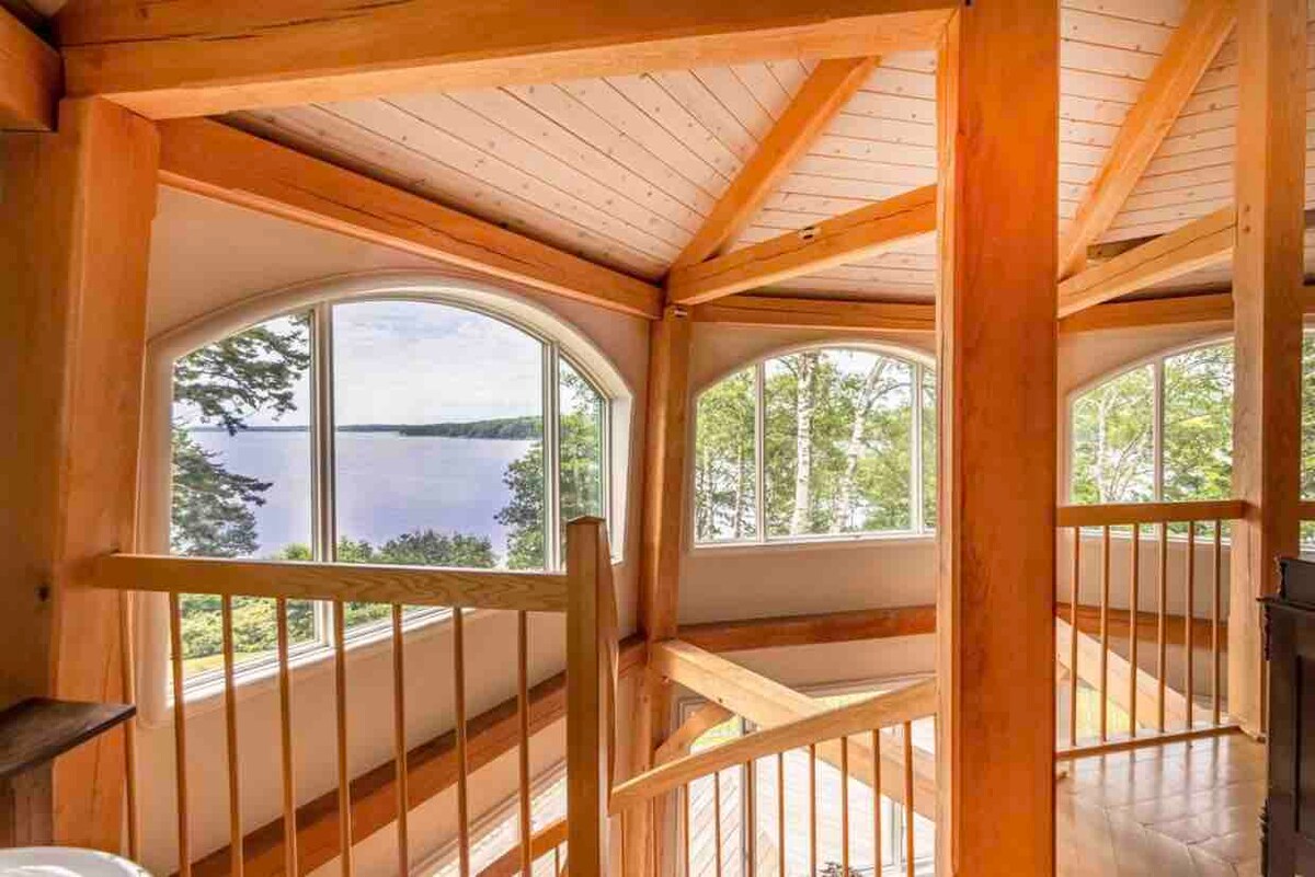 The image showcases the open loft space with vaulted wooden ceilings and large arched windows. Natural light floods the area, providing views of the serene waterfront. Wooden railings add a warm touch to the architecture, enhancing the connection to the surrounding nature.
