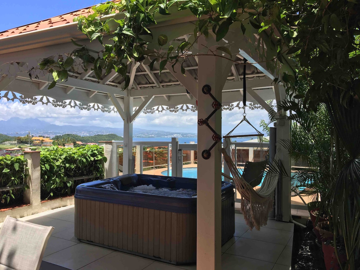 A shaded gazebo features a hot tub surrounded by lush greenery, offering a view of the Caribbean Sea. A hammock hangs nearby, providing a comfortable spot for relaxation. The layout encourages outdoor enjoyment with easy access to the pool area, creating a serene retreat.