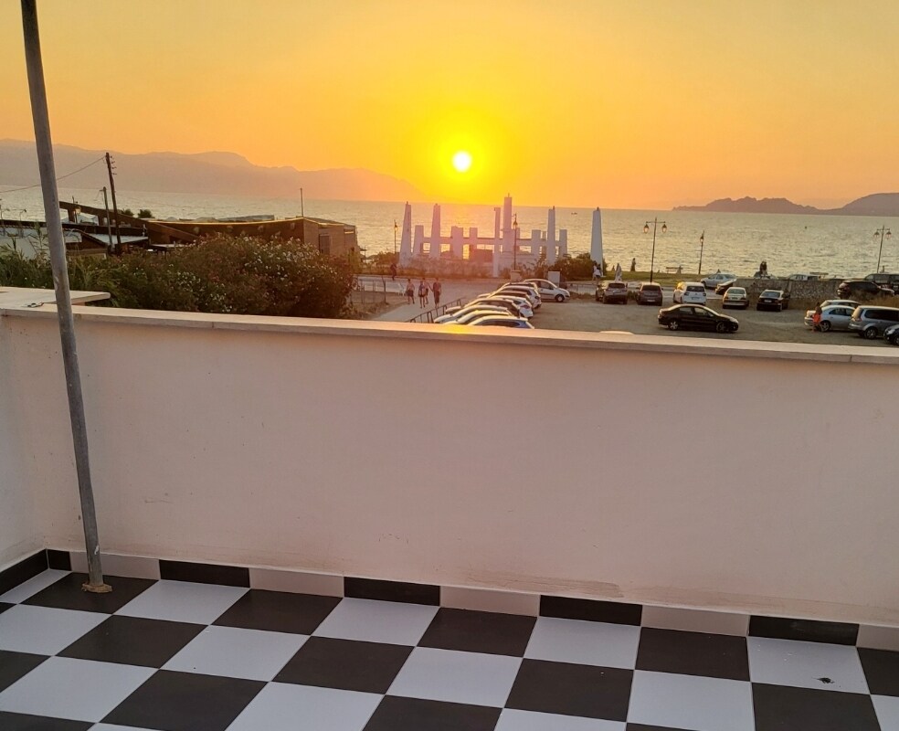 A sunset view is captured from a terrace, showcasing golden hues blending into the sky above the sea. In the foreground, a checkered floor adds a modern touch, while distant vehicles are parked along the shoreline, with silhouettes of people enjoying the view.
