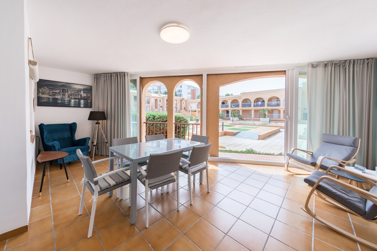 A bright living area is shown with tiled floors and large glass windows offering views of the shared garden. A dining table with six chairs is positioned centrally, accompanied by a comfortable blue armchair and a rocking chair in the corner.