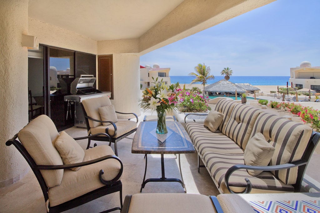 A spacious outdoor patio features comfortable seating, centered around a glass-topped table. The area is surrounded by blooming flowers and offers clear views of the ocean. A gas barbecue grill is visible nearby, enhancing the outdoor dining experience.