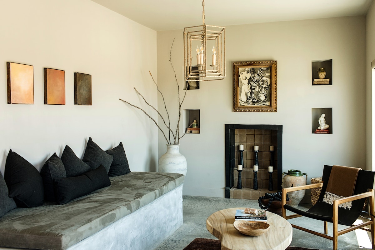 A spacious living room area features a built-in seating arrangement with plush, dark cushions against a wall. A decorative light fixture hangs above, illuminating the neutral-toned walls. A fireplace with candles is visible, complemented by an artistic wall display and a stylish coffee table.