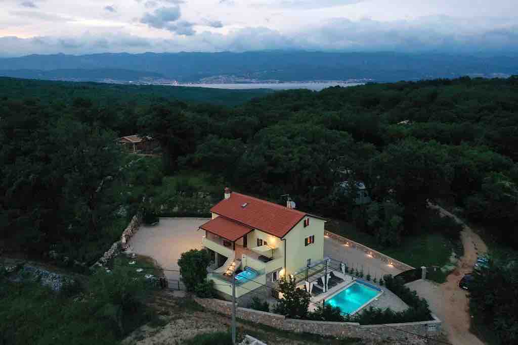 A scenic view of a two-story house set amidst lush greenery, featuring a charming red-tiled roof. A tranquil pool is positioned beside the house, with soft outdoor lighting enhancing the surroundings. The distant mountains and blue waters are visible under a cloudy sky.
