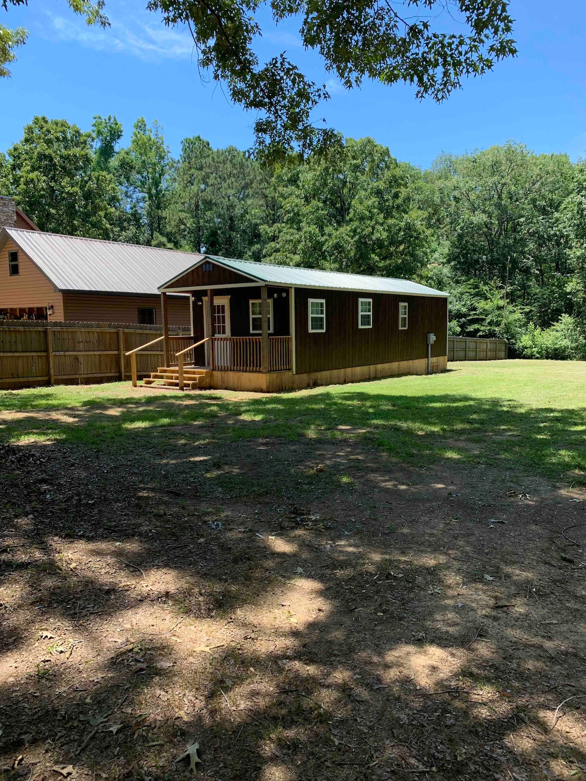 A small wooden house with a porch is set in a spacious grassy area, surrounded by trees. The building features a peaked roof and is situated away from neighboring houses, offering a sense of privacy. Sunlight filters through the leaves, creating dappled light on the ground.