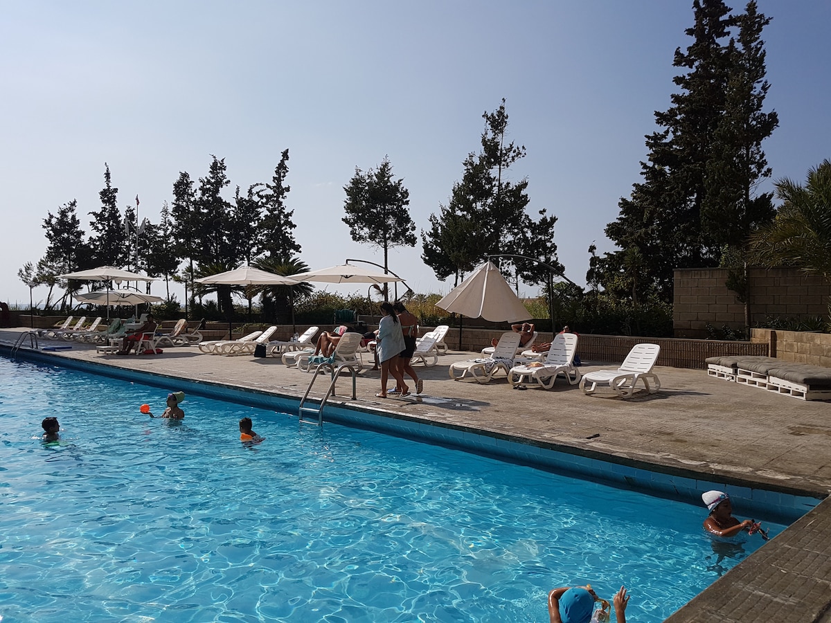 A spacious outdoor pool is surrounded by sun loungers and shade umbrellas. Lush trees line the background, providing a natural setting. A few guests enjoy the water, while others relax poolside, contributing to a lively yet tranquil atmosphere.