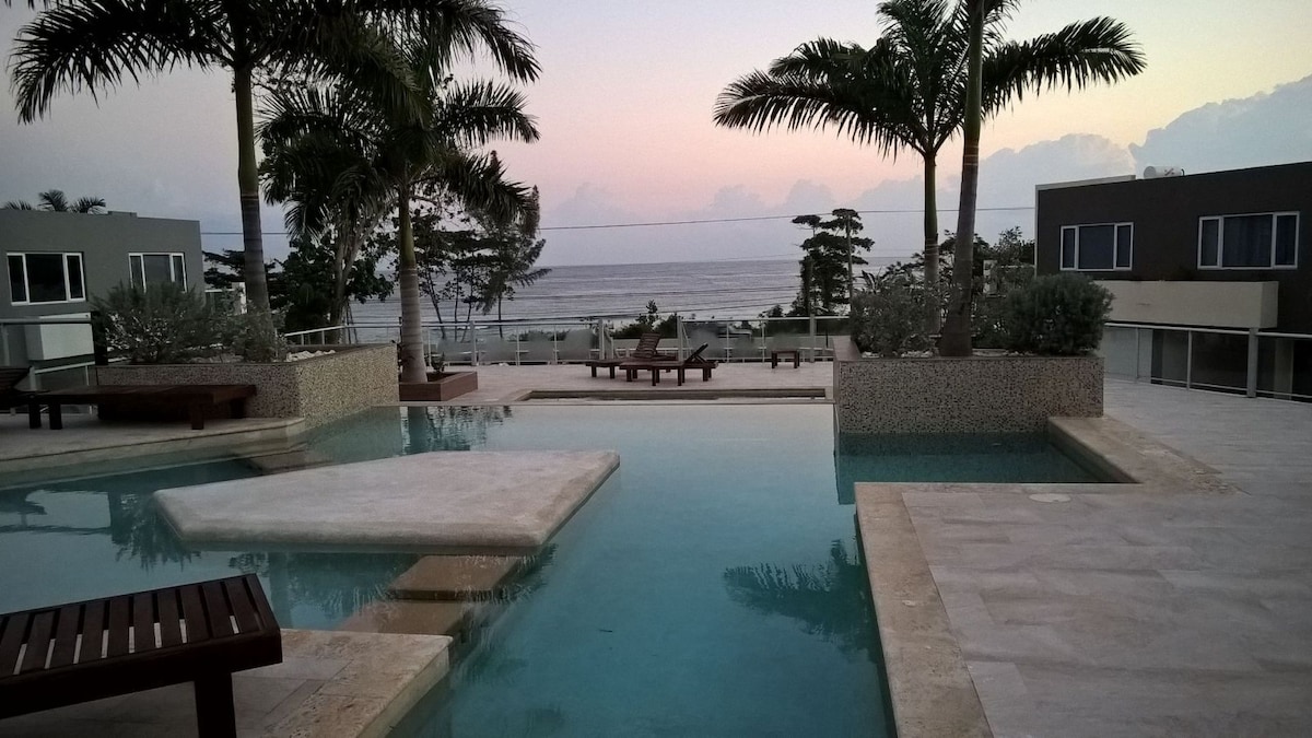 A serene infinity edge pool is presented, seamlessly flowing into the horizon where the ocean meets the sky. Lush palm trees surround the area, while wooden lounge chairs are positioned on a patio overlooking the water. Soft evening light casts a calming glow on the scene.
