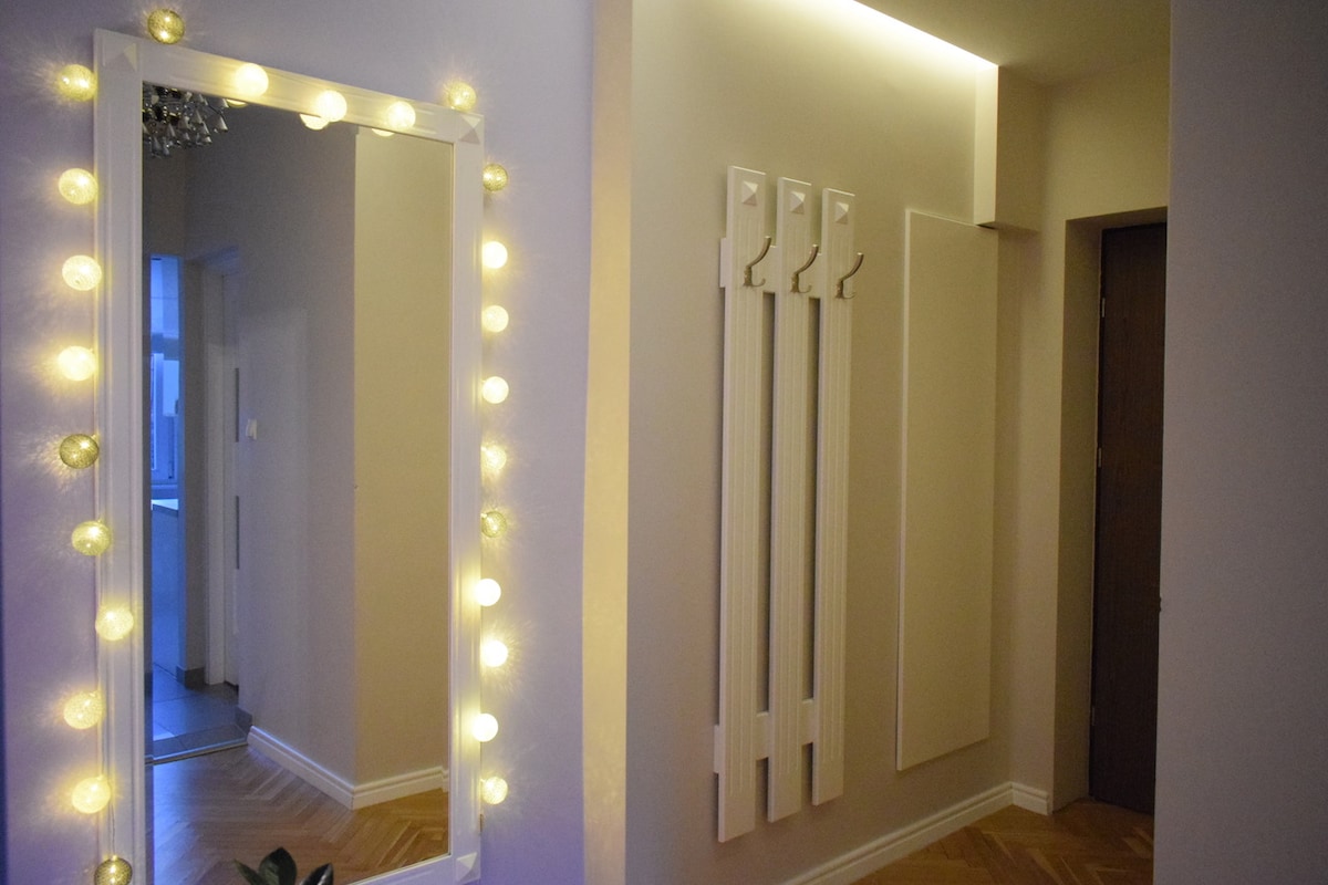 A well-lit entryway is shown, featuring a large mirror framed with soft lights. Four coat hooks are mounted on the wall beside the mirror. A door leads to another room, and warm wooden flooring enhances the inviting atmosphere.