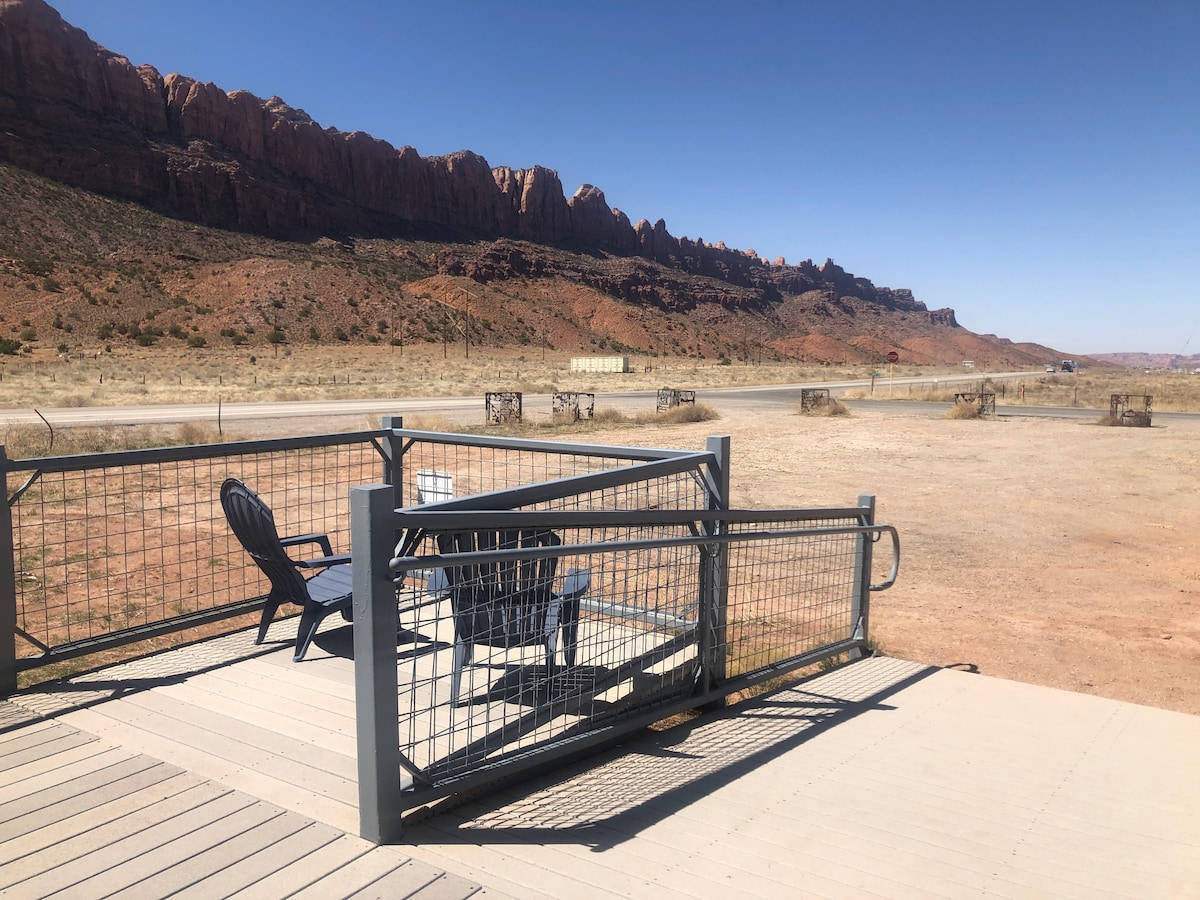 The deck features several chairs comfortably arranged, facing an expansive view of rugged red rock formations in the distance. The surrounding landscape is arid, with a gravel road visible nearby, offering a serene outdoor space for relaxation.