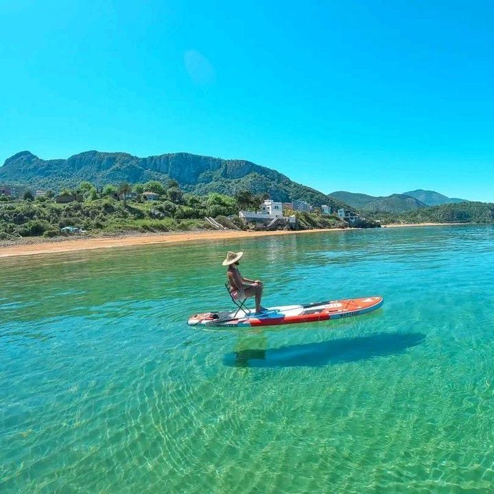 A calm water scene features an individual sitting on a paddleboard, surrounded by clear turquoise waters. Lush green mountains rise in the background under a bright blue sky. The shoreline, visible at a distance, is lined with a few houses.