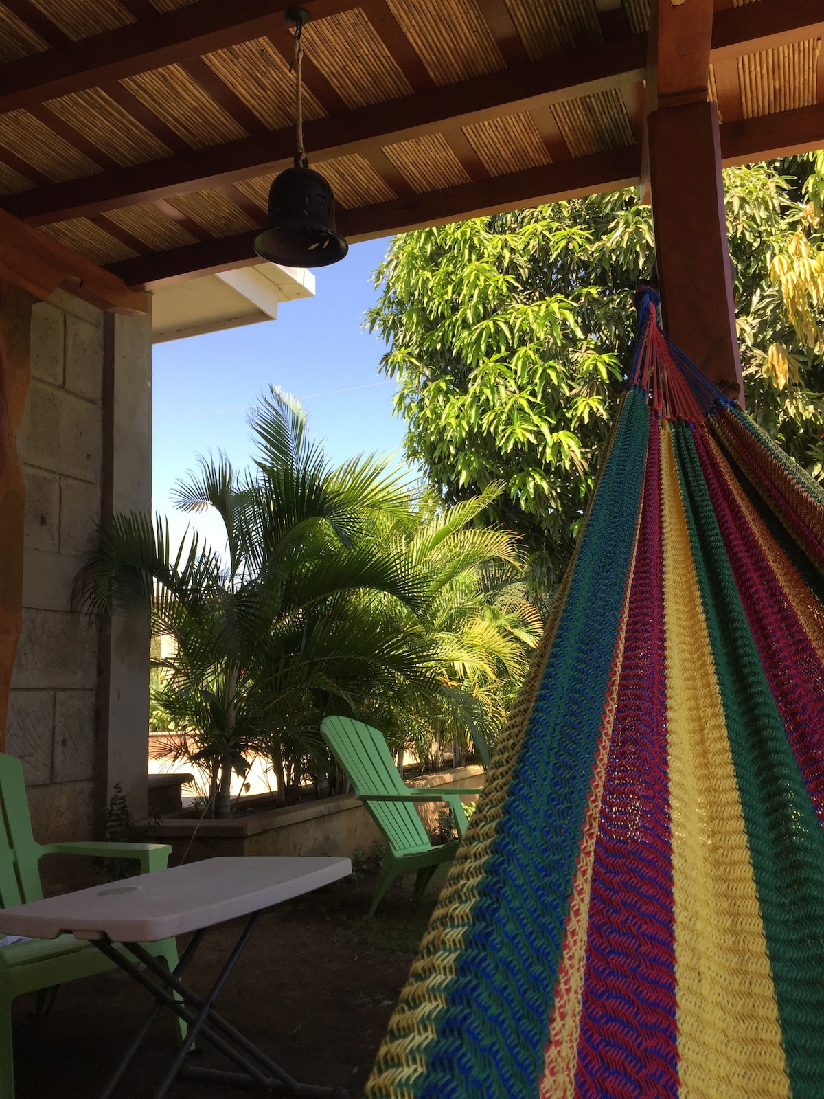 A colorful hammock hangs beneath a shaded patio, surrounded by lush greenery. Two light green chairs are positioned nearby, along with a small folding table. The clear blue sky peeks through the open space, creating a relaxing outdoor environment.