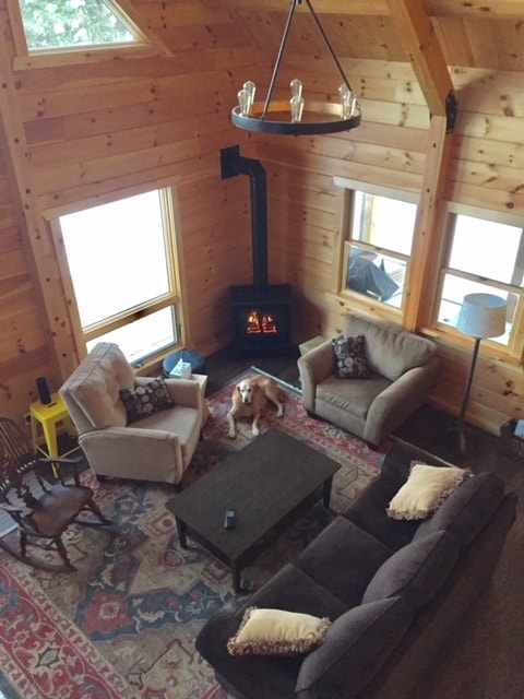 An open living area showcases natural wood walls and large windows allowing ample light. A cozy seating arrangement includes two armchairs, a sofa, and a coffee table on a patterned rug. A small lamp and a decorative chandelier add charm, while a dog relaxes near the wood stove.