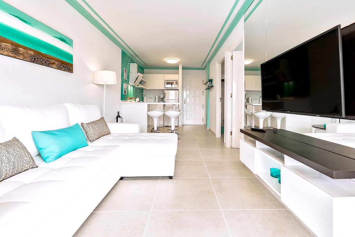 A modern living area is highlighted by a white sectional sofa adorned with turquoise and grey cushions. Light-colored tiles cover the floor, while a large flat-screen TV is mounted on the wall opposite a sleek console. The open kitchen area is visible in the background.