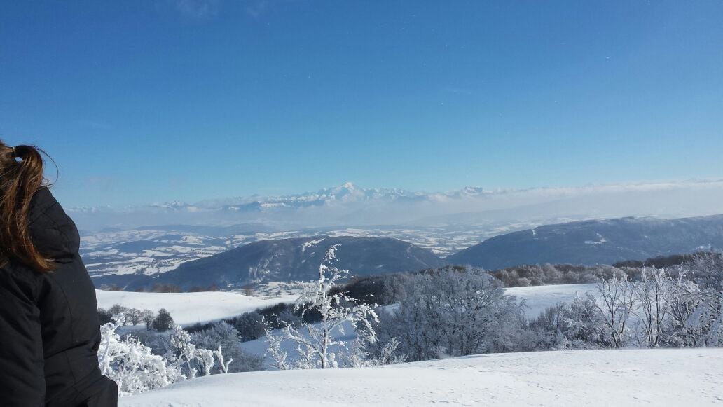 A snow-covered landscape is showcased, with rolling hills and trees blanketed in white. The distant mountains are partially visible against a clear blue sky, creating a serene atmosphere. The icy terrain reflects the sunlight, highlighting the beauty of the winter scenery.