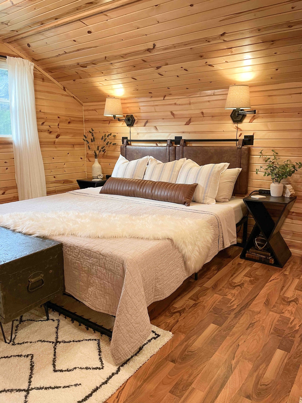 A cozy sleeping area features a king bed adorned with decorative pillows and a plush throw. The wooden walls provide a warm ambiance, while bedside sconces offer soft lighting. A small table holds a decorative plant, adding a touch of nature to the space.