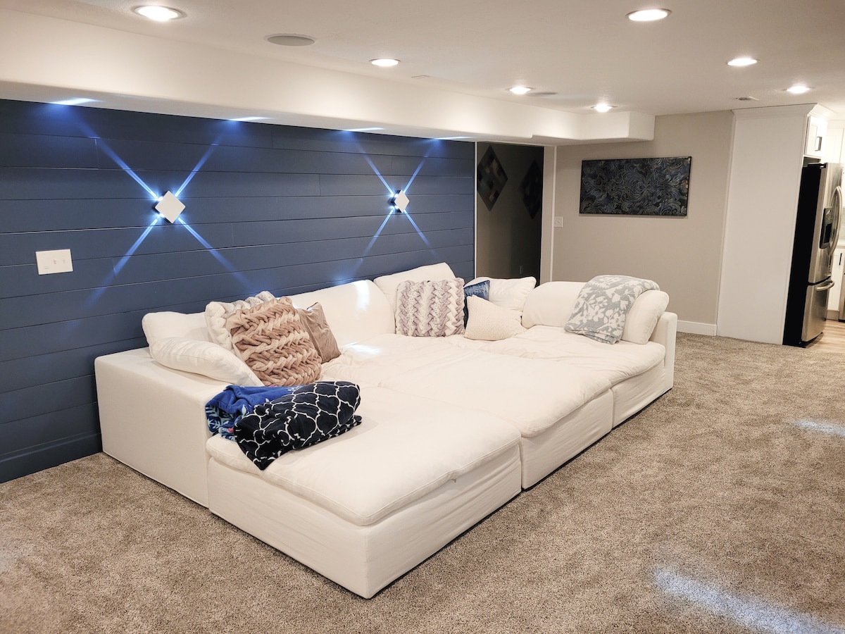 A spacious sectional sofa occupies the family room, featuring white cushions and a variety of decorative pillows. Soft lighting shines from the ceiling, complementing the blue accent wall. Plush carpeting covers the floor, creating a cozy atmosphere in the modern space.