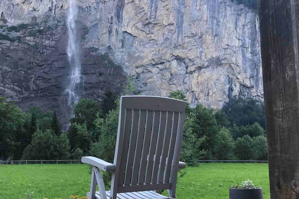 A sturdy wooden chair is positioned on a lush green lawn, providing a clear view of the dramatic cliffs and Staubbach Falls. Tall trees frame the scene, while vibrant flowers add a touch of color to the foreground.