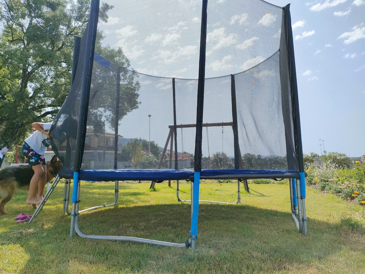 A trampoline is positioned in a green lawn area, surrounded by a protective net. A swing set is visible in the background, accompanied by trees and a partially cloudy blue sky.