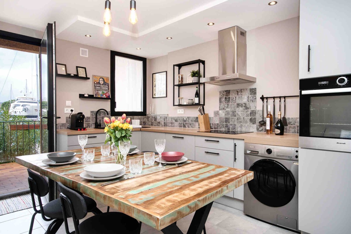 A modern kitchen features minimalist cabinetry and stylish countertops. A dining table made of wood provides seating for six, complemented by elegant tableware. A selection of kitchen appliances is neatly arranged, with natural light streaming in from a nearby window overlooking the marina.