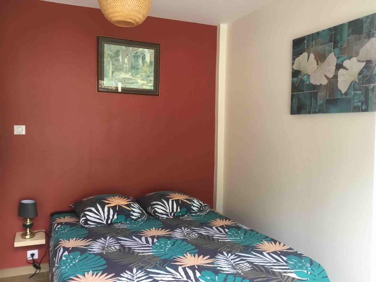 A serene bedroom features a double bed adorned with botanical-patterned bedding. A subtle wall painting adds character to a terracotta accent wall. A bedside lamp is positioned on a wooden nightstand, contributing to a calm and restful environment.