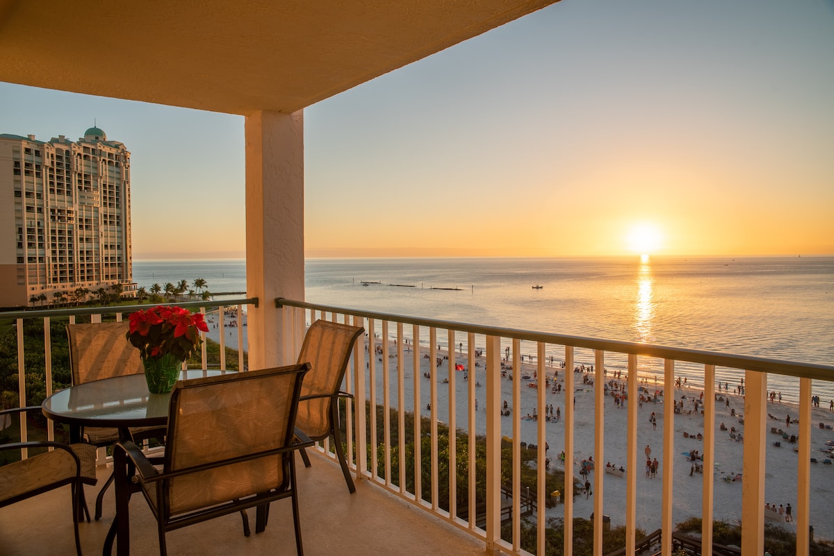 A private balcony provides an unobstructed view of the sunset over the Gulf. A table with four chairs is positioned for outdoor dining, while colorful flowers add a touch of vibrancy. The beach and distant skyline are visible, creating a serene coastal atmosphere.
