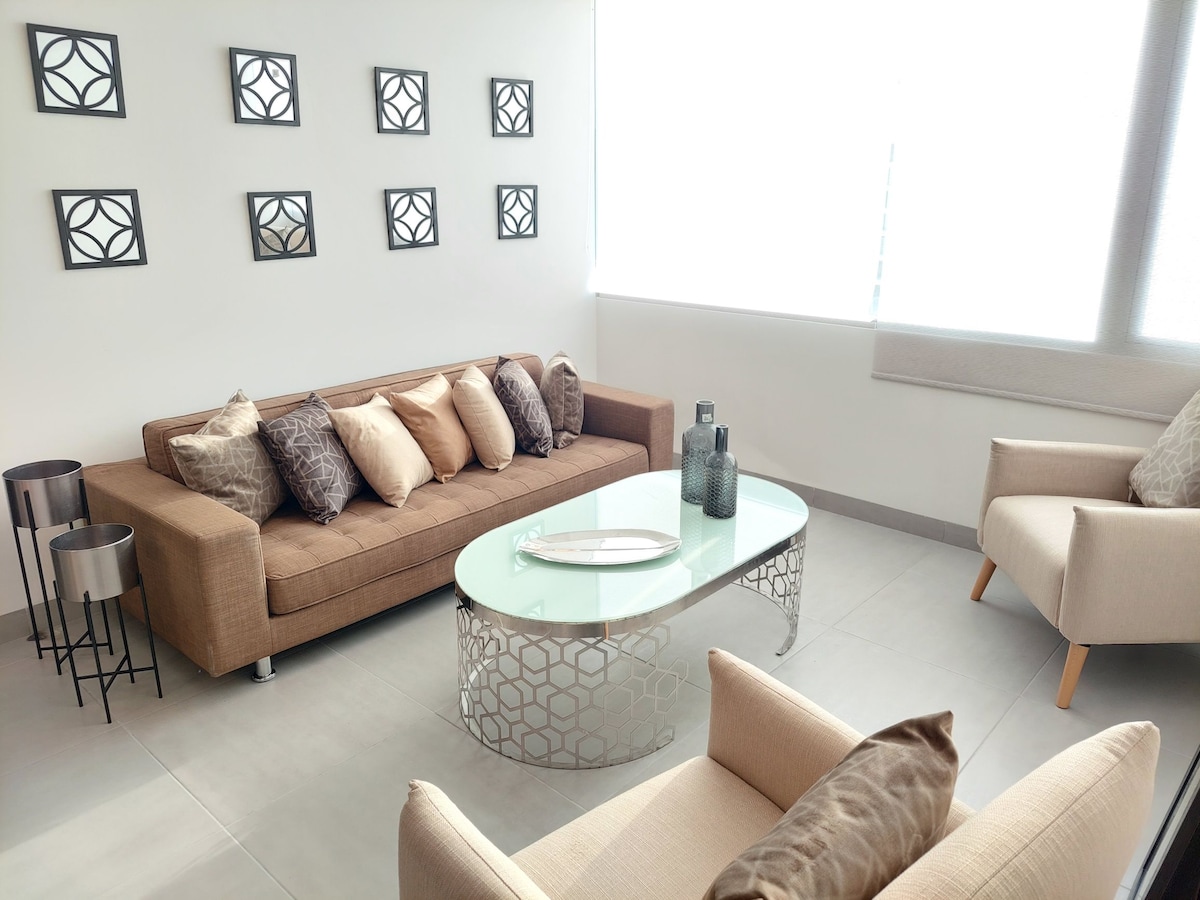 A light-filled living area features a modern brown sofa adorned with decorative cushions, complemented by two beige armchairs. A glass coffee table with a textured base sits centrally, enhancing the clean and spacious layout. Wall art consists of six framed pieces arranged in a grid.