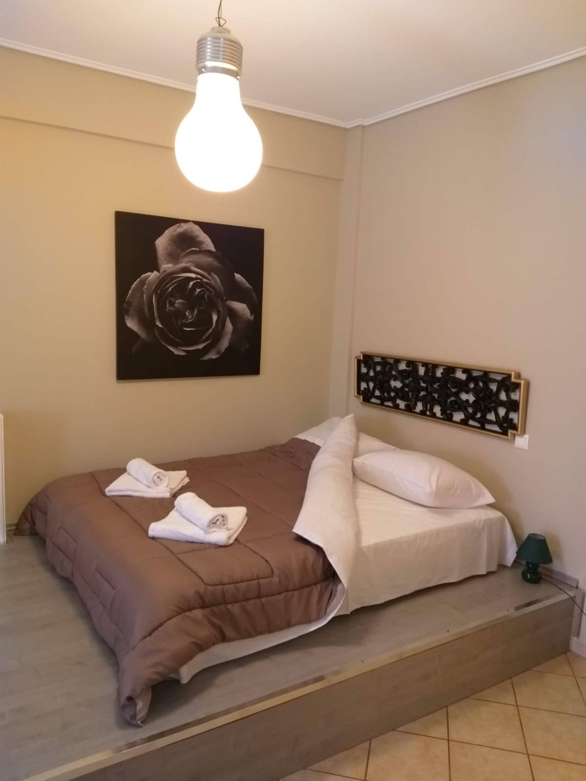 A spacious sleeping area features a raised bed dressed in neutral tones with rolled towels arranged neatly on the covers. A large black and white art piece of a rose hangs above the bed, accompanied by a minimalist headboard. Soft lighting is provided by an overhead bulb.