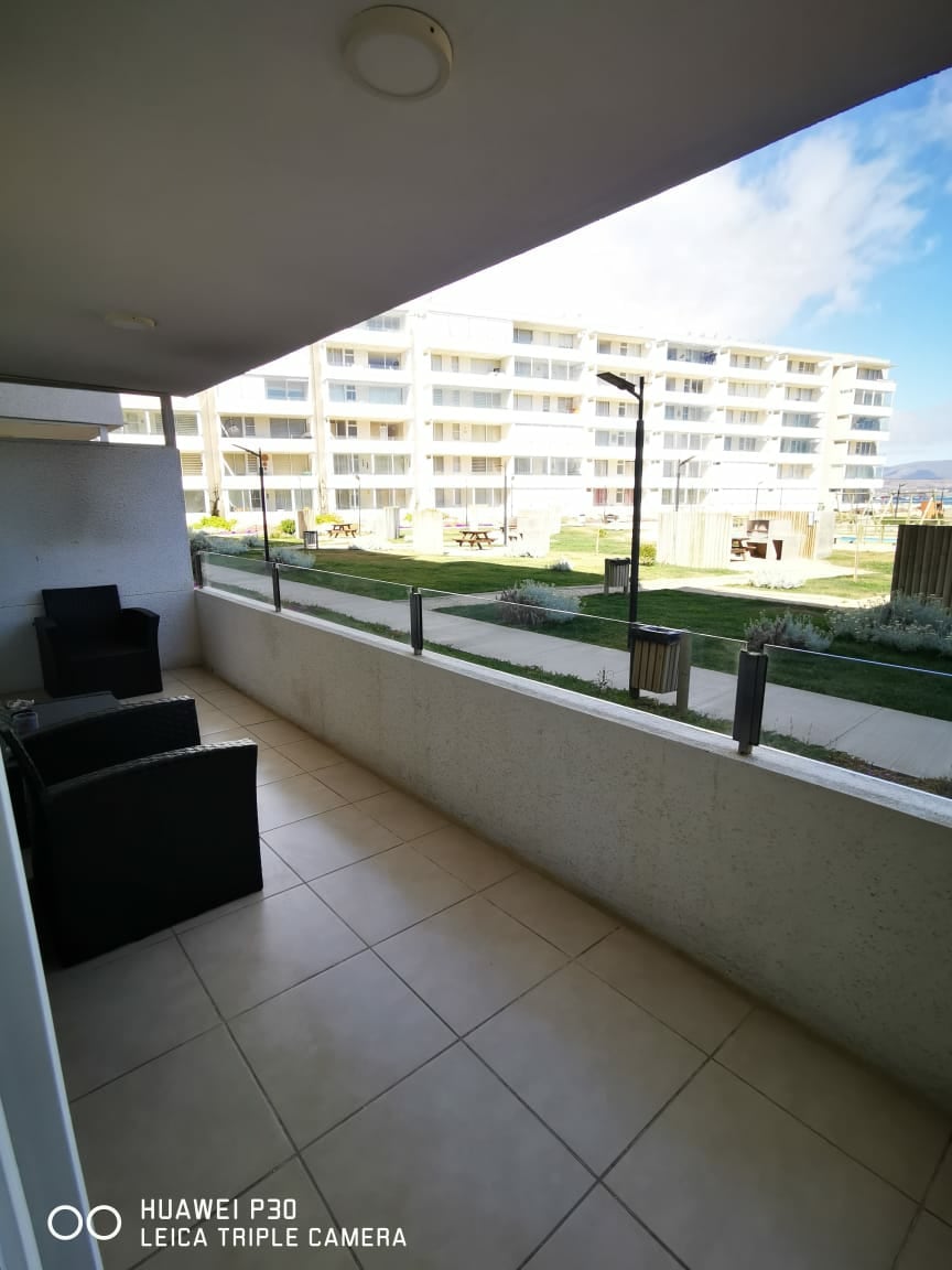 A private balcony features modern outdoor furniture, including two dark chairs and a small table. The view includes a green lawn with several seating areas and a well-maintained garden, framed by a contemporary apartment building in the background.