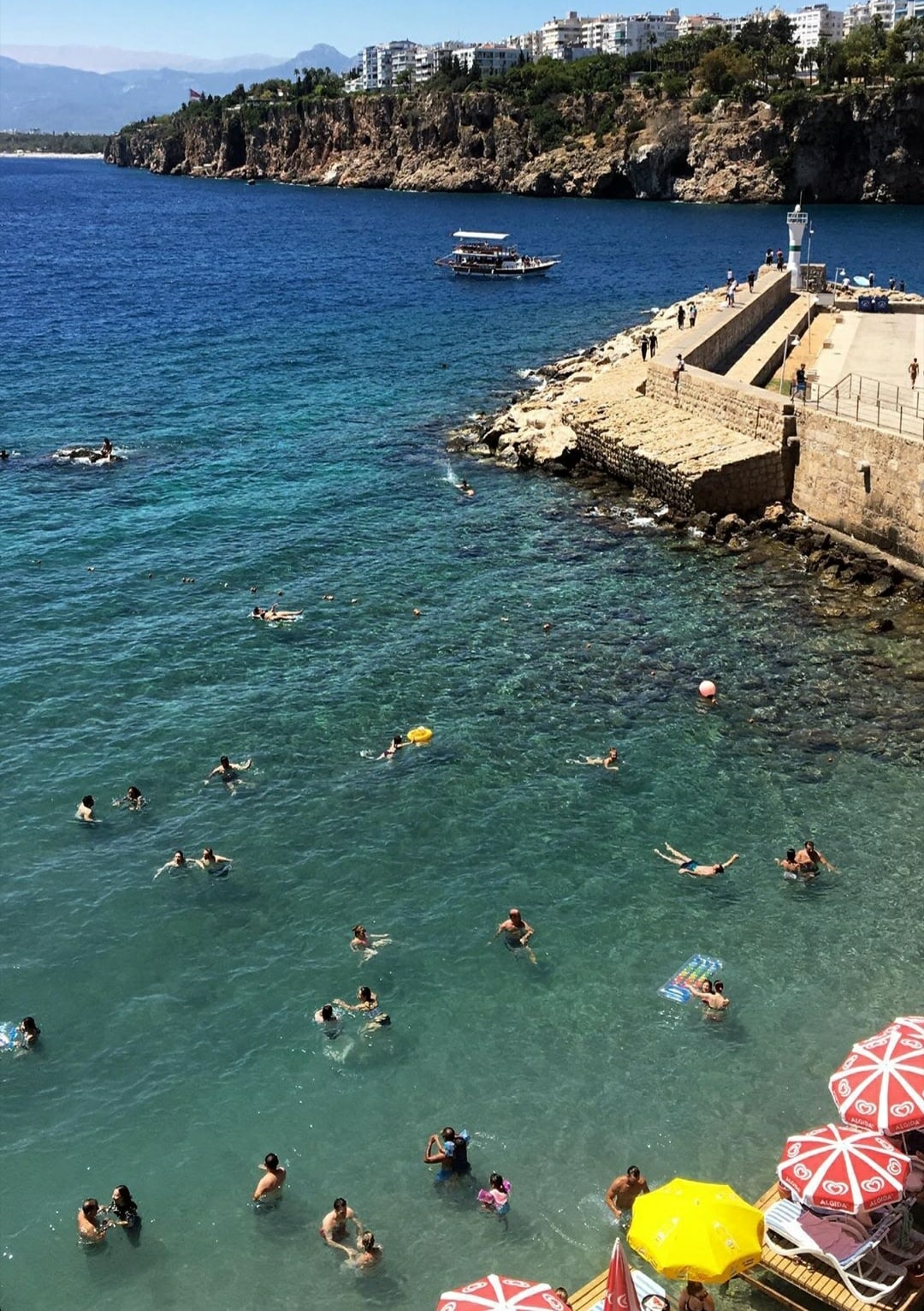 The image depicts a vibrant seaside scene featuring people swimming in clear blue waters. A rocky pier extends into the ocean, with a boat visible nearby. Beach umbrellas in various colors line the shore, adding to the lively atmosphere.