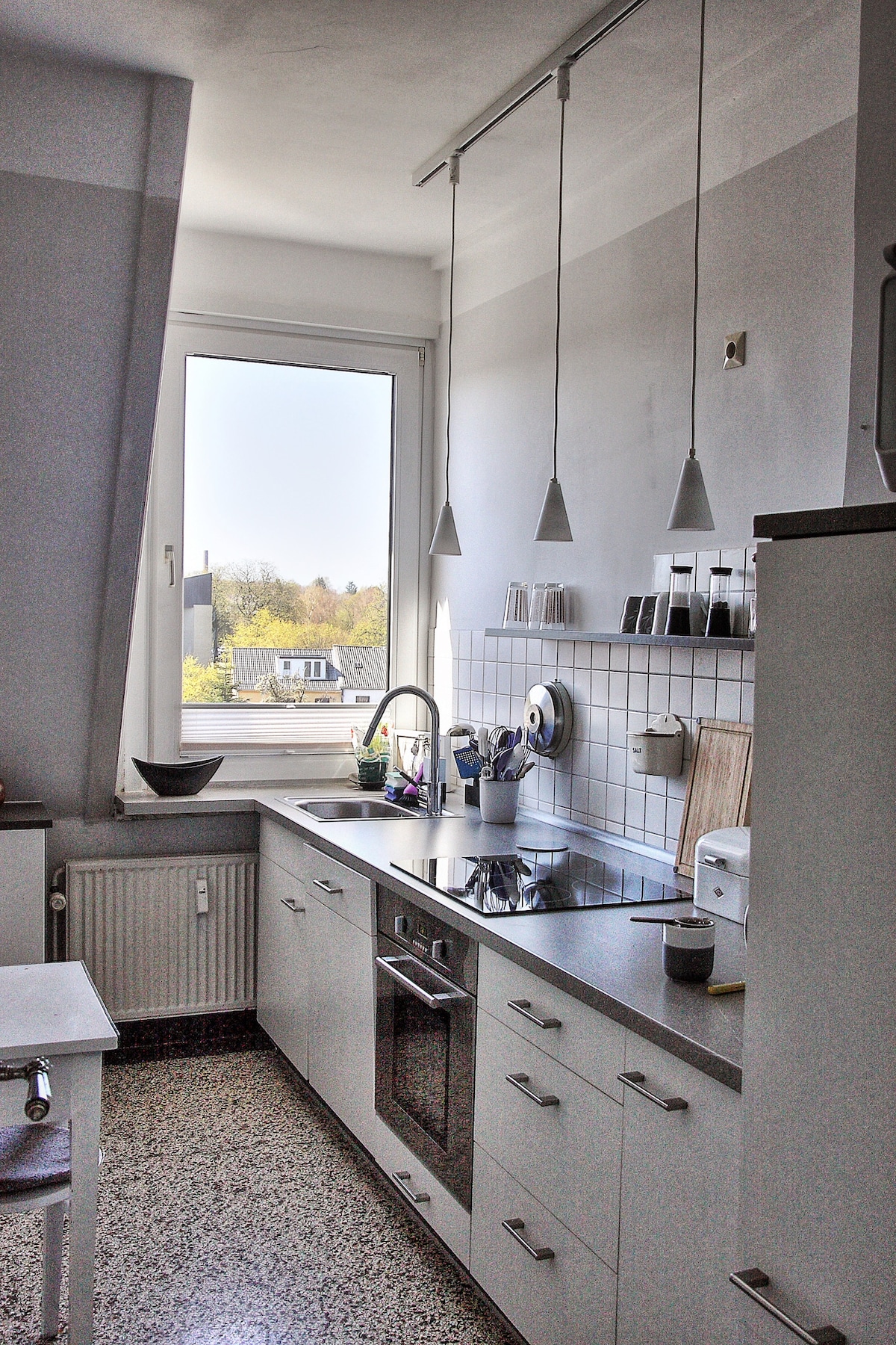 A modern kitchen features sleek cabinetry and a countertop with built-in appliances. Three pendant lights hang above the workspace, which is illuminated by natural light from the large window overlooking the city. Minimalistic decor is complemented by organized shelves of glassware.