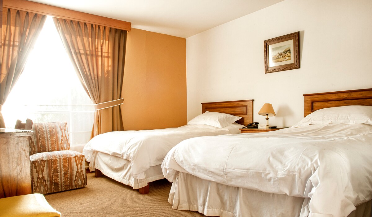 A well-lit guest room features two spacious beds with white linens and wooden headboards. Sunlight filters through curtains, casting a warm glow across the room. A small bedside table with a lamp is positioned between the beds, accompanied by a patterned chair nearby.