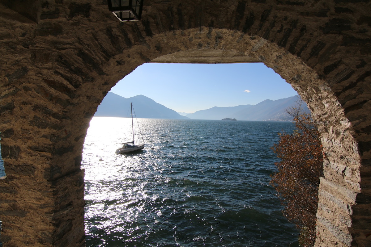 A scenic view is framed by an arched stone structure, showcasing a sailboat on the tranquil waters of Lago Maggiore. The sun reflects off the surface, creating shimmering patterns, while distant mountains provide a backdrop under a clear sky.