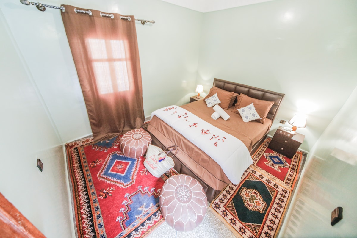 A well-appointed bedroom is showcased, featuring a spacious bed adorned with a brown and white coverlet. Decorative pillows add a touch of color. Two bedside tables with lamps flank the bed, while traditional rugs enhance the room's character. A dark curtain partially covers a window.