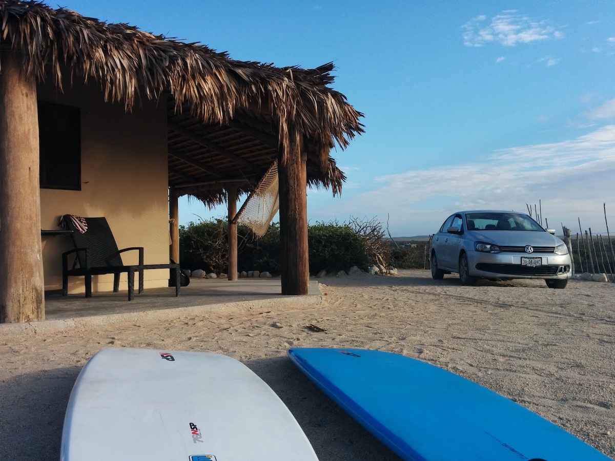 A rustic beach house is viewed from a sandy area, showcasing a thatched roof and wooden posts. Two surfboards rest on the ground, while a car is parked nearby. The sky is clear with a few clouds, enhancing the coastal ambiance.