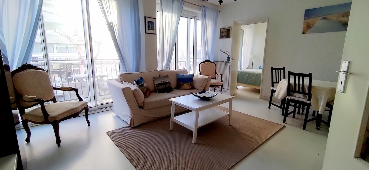 A bright living area features large double-glazed windows draped with light blue curtains, allowing natural light to fill the space. A spacious white sofa is positioned near a minimalist coffee table, complemented by two armchairs. A dining table and chairs are also visible in the background.