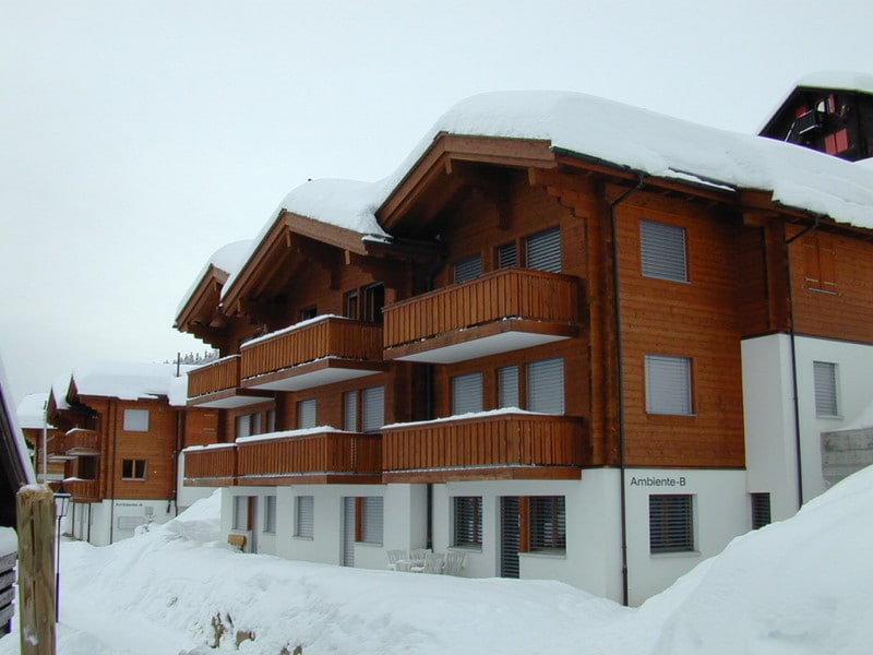 A wooden chalet-style building is visible, featuring multiple balconies adorned with wooden railings. The structure is partially covered in snow, highlighting its winter setting. Large windows are present, offering a glimpse of the cozy interior. The overall atmosphere is one of a serene mountain retreat.