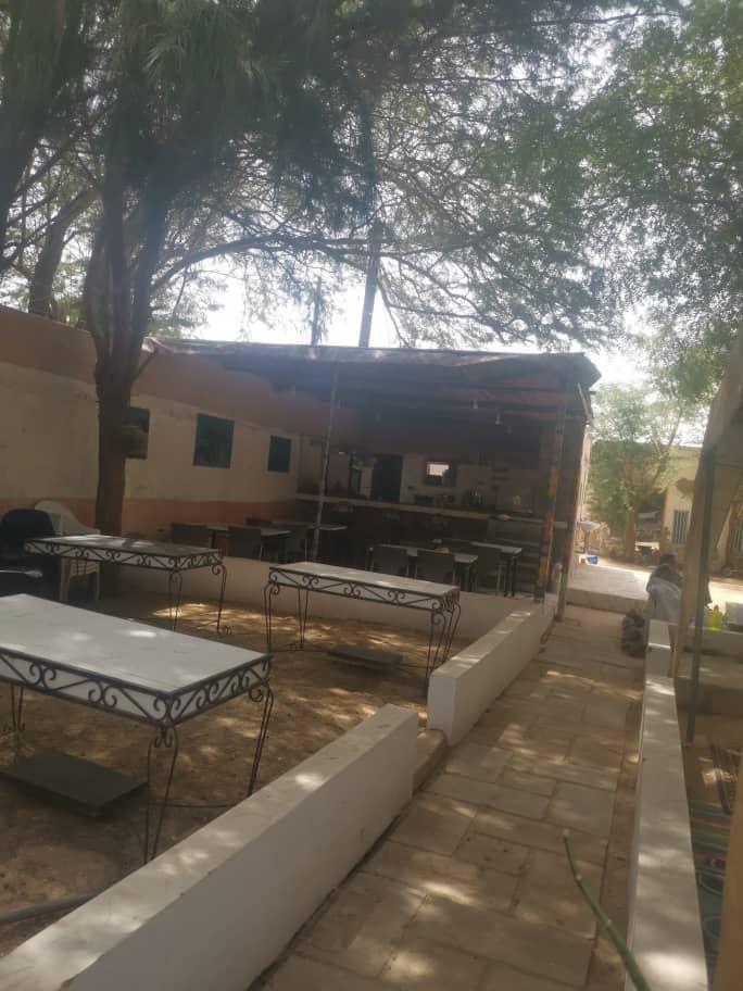 An outdoor seating area is surrounded by trees, featuring several tables with elegant metal frames. The pathway, composed of stone tiles, leads to a shaded structure where communal cooking and dining may take place. Soft sunlight filters through the trees, enhancing the peaceful ambiance.
