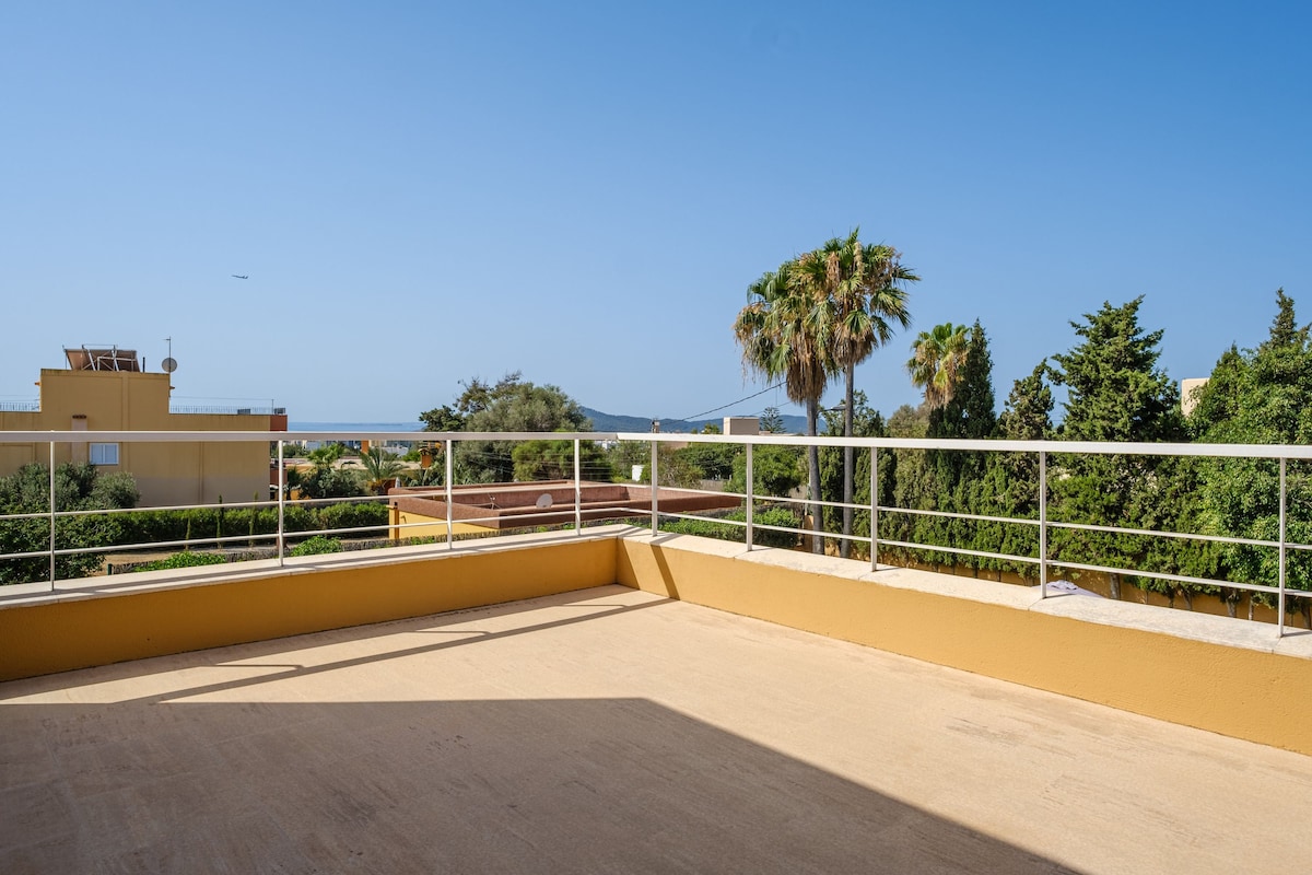 A spacious balcony is showcased, featuring a railing that offers an unobstructed view of lush greenery and the distant sea. The bright blue sky is visible above, enhancing the open atmosphere of the outdoor space.