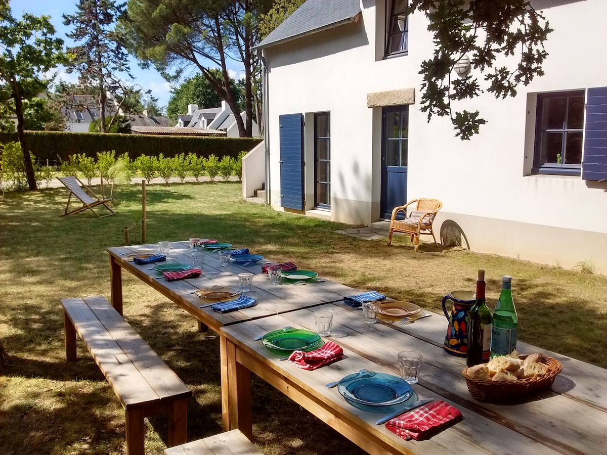 An outdoor dining area is set up on a wooden table, featuring colorful plates and glasses arranged for a meal. Nearby, a comfortable rattan chair and a sun lounger are visible on the grassy lawn, surrounded by trees and greenery.