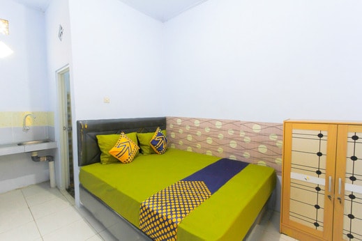 A well-appointed room features a spacious bed dressed in bright green linens and accented with decorative pillows. A small sink and mirror are visible in one corner, while a wooden cabinet provides storage. Soft light enters through a nearby window.