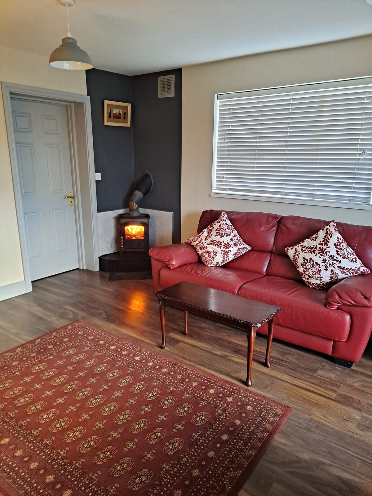 A living area features a red couch adorned with patterned cushions, positioned beside a small wooden coffee table. A decorative area rug covers the floor, and a wood-burning stove adds warmth to the corner. Natural light filters through the window with horizontal blinds.