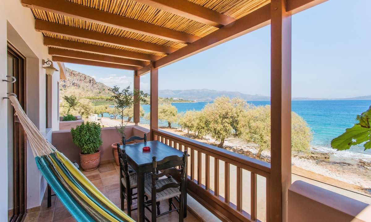 A balcony features a wooden table set for outdoor dining, complemented by a colorful hammock. Lush greenery contrasts with the blue sea visible in the background, offering a serene coastal view. The roof provides shade, enhancing the inviting atmosphere of this outdoor space.