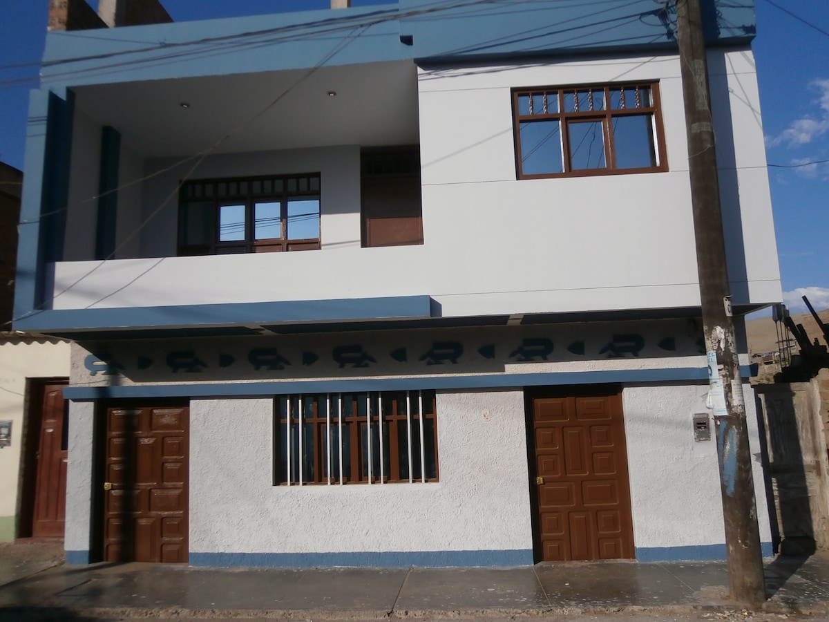 The exterior of a two-story building is displayed, featuring a combination of white and blue walls. The upper floor has large windows with wooden frames, while the lower level has two wooden doors. The structure is located along a modest street.