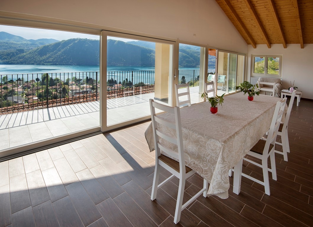 A spacious dining area is illuminated by natural light, featuring a large table surrounded by white chairs. Floor-to-ceiling glass doors offer expansive views of the lake and surrounding landscape, enhancing the connection to the outdoors.