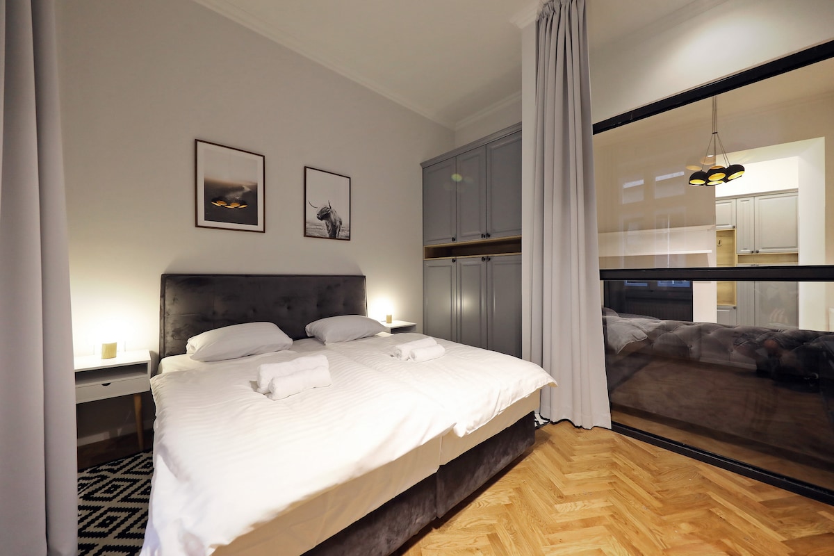 A neutral-toned bedroom features two spacious beds with white linens, complemented by two bedside tables with lamps. Framed artwork adorns the walls. A glass partition leads to a visible kitchen area, enhancing the open concept design. Natural light filters through the curtains.