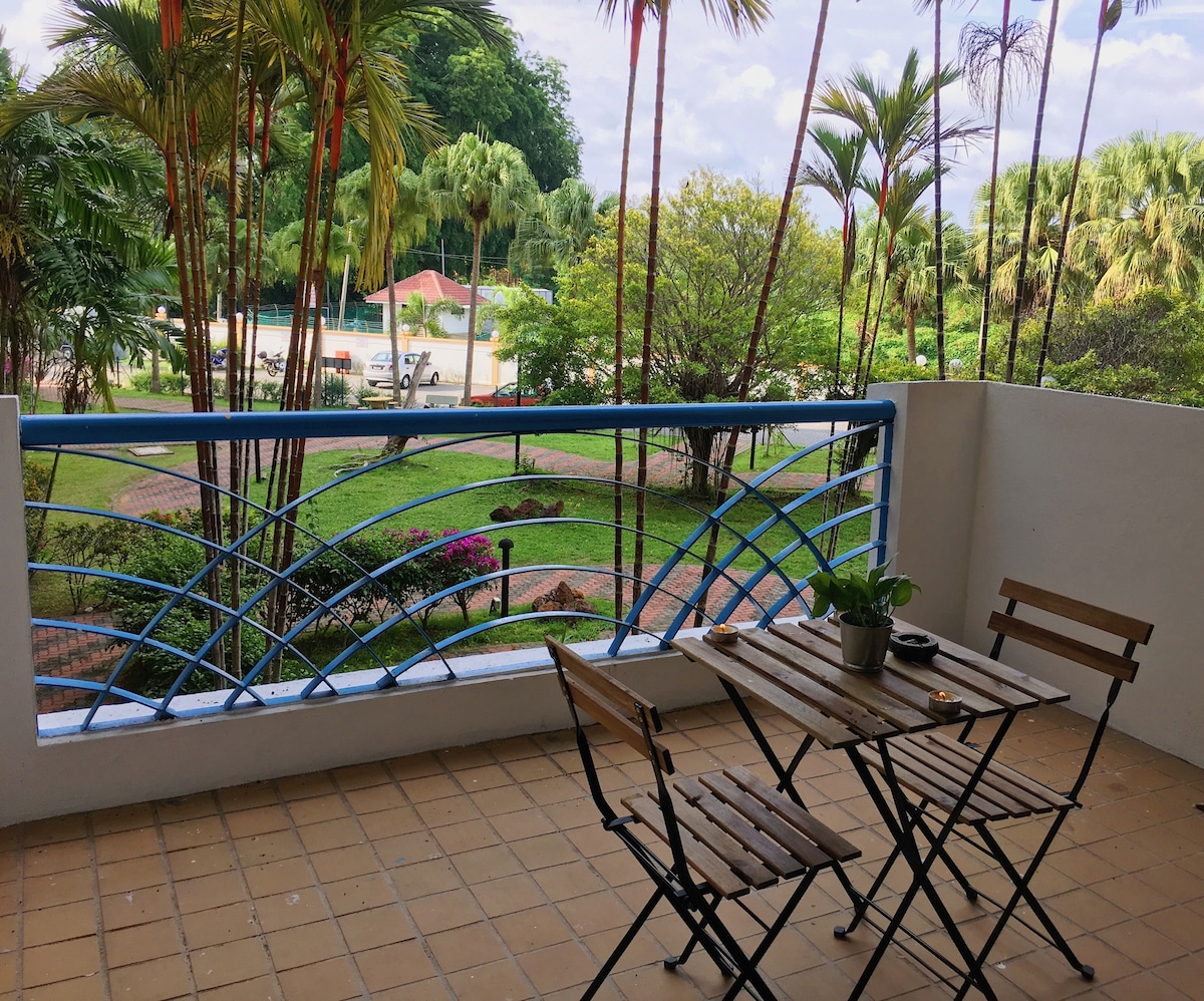 A balcony area features a small wooden table and two chairs, surrounded by lush greenery and vibrant landscaping. Palm trees and flowering plants create a serene outdoor atmosphere, with the view extending to well-maintained grounds.