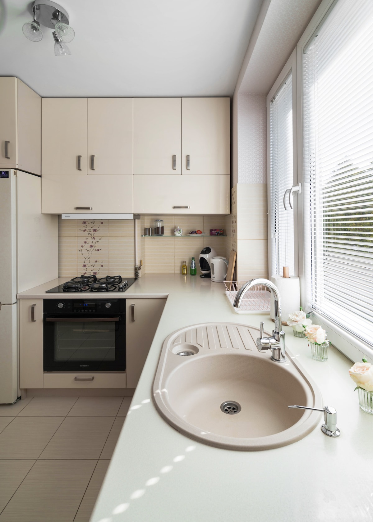 A modern kitchen features light-colored cabinetry and a sleek countertop. An oven and gas stove are integrated into the design, along with a sink that has a contemporary faucet. Natural light streams through large windows, illuminating the space and highlighting decorative elements.