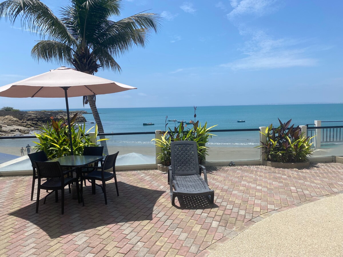 An outdoor terrace features a round dining table with four chairs under a large umbrella. A lounge chair is positioned nearby. Lush greenery surrounds the area, offering a view of the calm sea with a few boats visible in the distance.