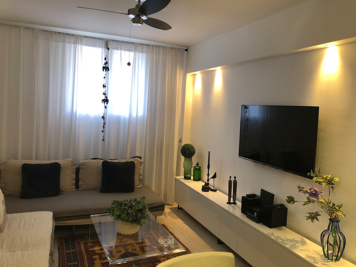 A light-filled living area is displayed with a comfortable seating arrangement featuring neutral-toned cushions. A large wall-mounted television is positioned across from a minimalist console. Natural light filters through sheer curtains, enhancing the space's inviting ambiance.