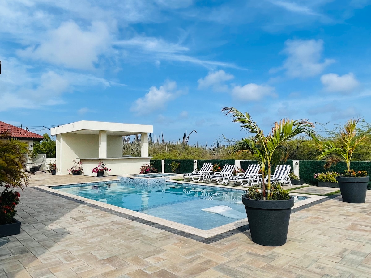 The outdoor area features a spacious pool surrounded by a stone patio, with several lounge chairs positioned for relaxation. Tropical plants are displayed in large pots, and a shaded bar area is seen in the background, creating a comfortable space for outdoor enjoyment.